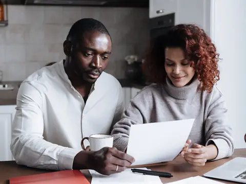A couple sitting at a kitchen table calmly reviewing a sheet of paper, with coffee mugs, a pen, documents and a laptop in front of them
