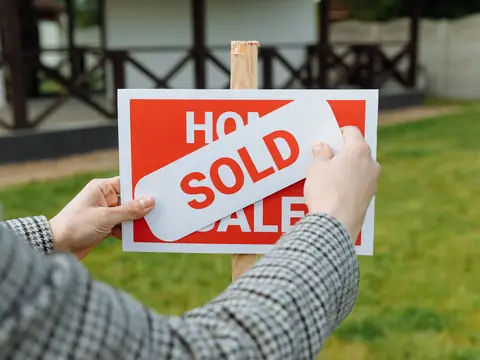 Two hands pressing a white SOLD sticker across a red and white FOR SALE yard sign, with a green lawn and a house blurred behind